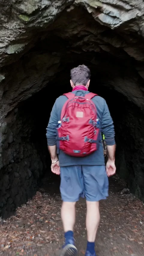 Man with red backpack walks into a dark cave tunnel, Karekare, New Zealand 動画素材 335600557