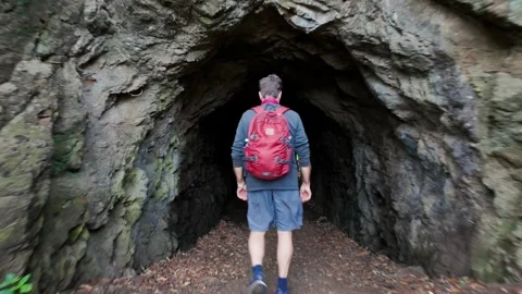 Man with red backpack walks into a dark cave tunnel, Karekare, New Zealand 動画素材 335601852