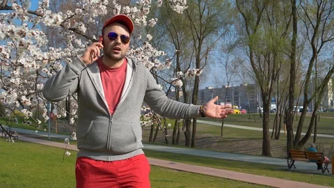 Man in red baseball cap in sunglasses talks on smartphone in city park Vídeo Stock 110842526