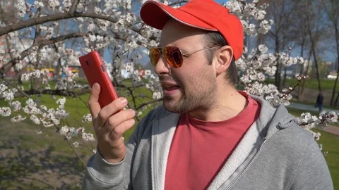 Man in red baseball cap in sunglasses talks on smartphone in city park Vídeo Stock 114283358