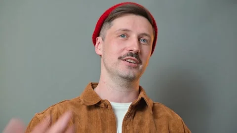 Man with a red beanie and brown jacket speaks and smiles in a studio setting. Видео 337642585