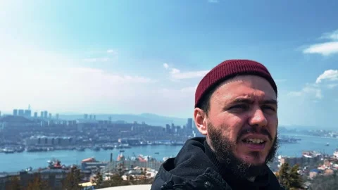 A man with a red beanie on his head is standing on a hill overlooking a city Stock Footage 308920287