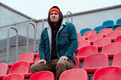 Man in a Red Beanie Sits on Empty Stadium Seats During a Chilly Day in a Qu.. Stock Photos