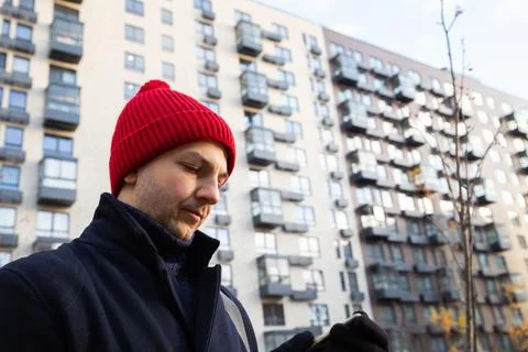 Man in red beanie using mobile phone outdoors near apartment building Foto stock