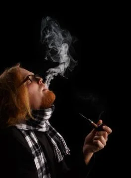 Man with red beard in chequered scarf and glasses holds tobacco pipe and pu.. Stock Photos
