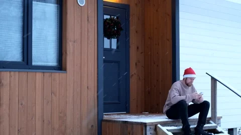 A man in a red cap with a phone sits on the porch of a house in snowy weather Stock Footage 295154270