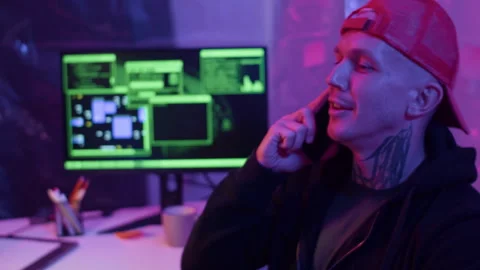 Man in Red Cap Talking on Smartphone in Neon-Lit Room with Code on Monitor Stock Footage 303561439