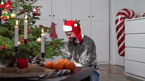A man in a red christmas hat puts a gift under a christmas tree in a cozy home Stock Footage 167514947