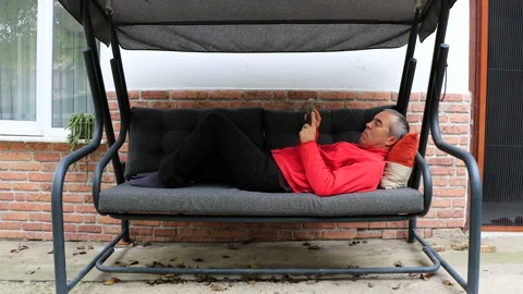 A man in red clothes is lying on a swinging sofa in the garden Vídeos de archivo 290029991