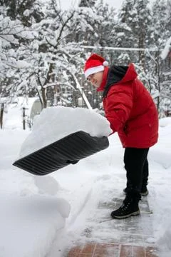 Man in red down jacket and red hat of Santa claus clears snow in backyard. Cl Stock Photos