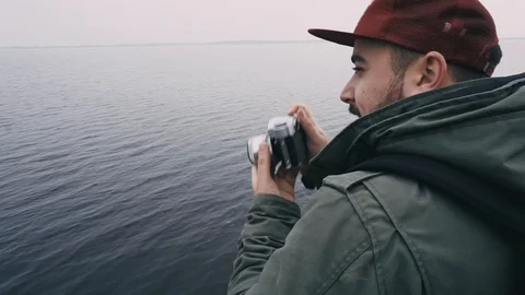 Man in the red hat with dark beard making a shot with camera on the sea shore Stock Footage 97792118