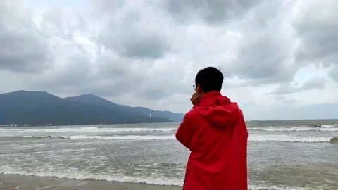 Man in red hoodie in a cold cloudy day by the wavy blue sea Stock Footage 146251332