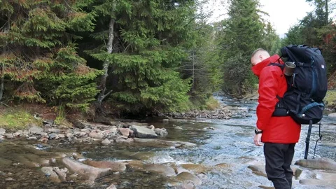 A man in a red jacket and with a black tourist backpack near a mountain river 動画素材 120236470