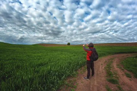A man in a red jacket with a backpack walks along a dirt road in a green field Stock Photos