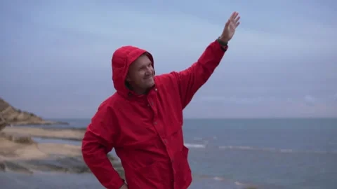 Man in red jacket on beach, waving hand with water and sky in background Stock Footage 274032769