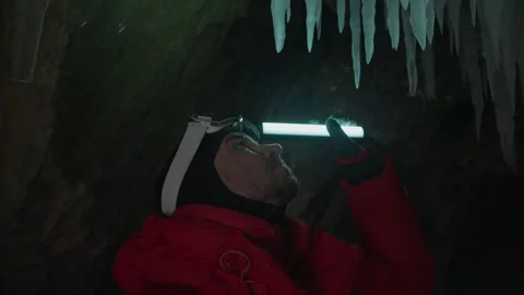 Man in red jacket with led light watch up, illuminate icicles hangi in ice cave Stock Footage 166566479