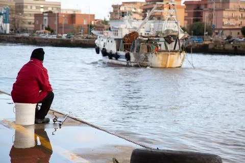 A man in a red jacket sits on a dock, watching a boat Stock Photos