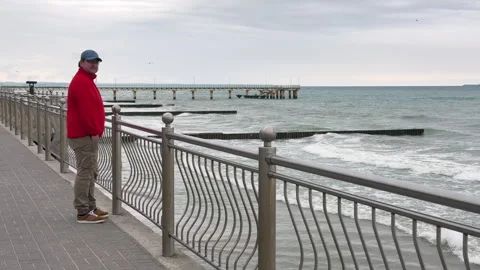 A man in a red jacket stands on the Baltic Sea promenade on a cloudy day Stock Footage 310682299