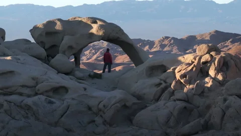 A man in a red jacket stands by a natural rock arch in the desert landscape Stock Footage 274477867
