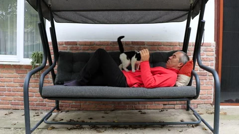A man in red is lying on a swinging sofa in the garden of his house with his cat Vídeos de archivo 290024797