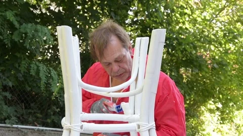 Man in red protective coverall painting chair outdoors during home renovation wo Stock Footage 327654341