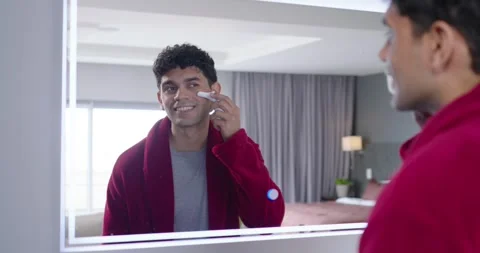 Man in red robe checking reflection, grooming jawline at mirror with electric Stock Footage 348223575