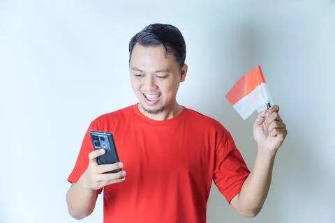 A man in a red shirt smiling while looking at his phone, holding an Indones.. Stock Photos