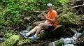 Man with red smartphone sits on a stone near small river and listens to music HD Footage