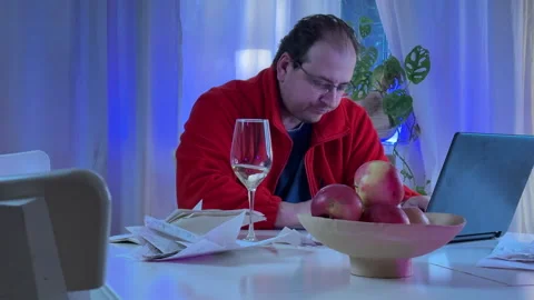 A man in a red sweater and glasses sits at a white kitchen table in the evening  Vídeo Stock 306014273
