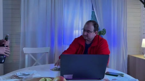 A man in a red sweater and glasses sits at a white kitchen table in the evening  스톡 동영상 306014936