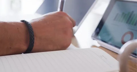 Man referencing tablet charts and writing notes at desk with pen in lined 動画素材 344949577