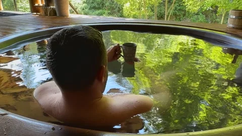 A man relaxes in a hot bath, drinks tea from a cup, enjoying the sunlight and Stock Footage 251747024