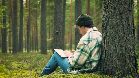 Man relaxes in a peaceful forest, reading a book and drinking from a flask. Outd Stock Footage 344405626