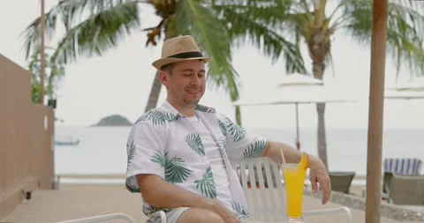 Man relaxing beachside in tropical setting, wearing hat and floral Stock Footage 314046367