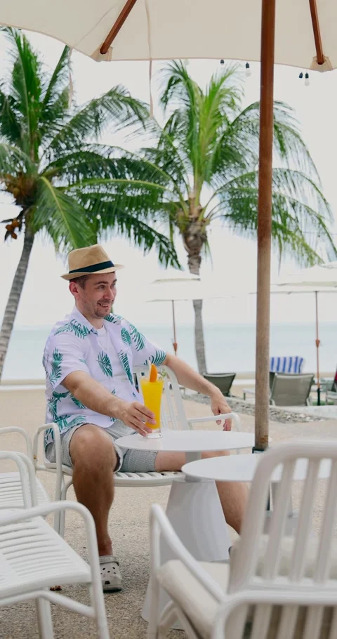 Man relaxing beachside under umbrella holding tropical drink, palm Stock Footage 314045791