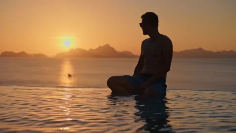 Man relaxing on the edge of luxury infinity pool, sunset view reflected in water Video stock 282905709