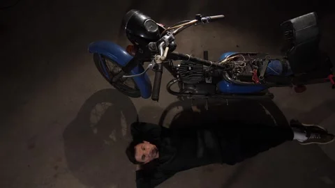 Man relaxing on garage floor, looking up at a disassembled 1970s blue Stock Footage 342767234