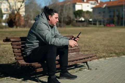 Man relaxing on park bench using smartphone and smoking Foto stock