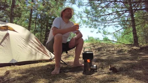 Man Relaxing with Tea by Tent in Pine Forest, Kettle on Wood Stove Stock Footage 281630733