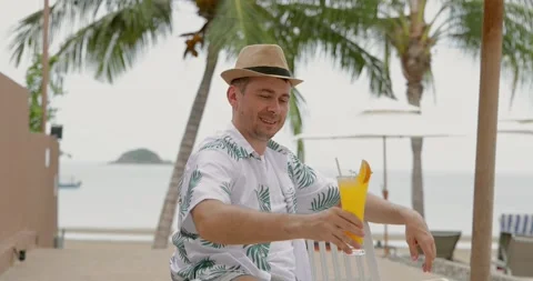 Man relaxing in tropical beach setting, wearing straw hat and floral Видео 314047158