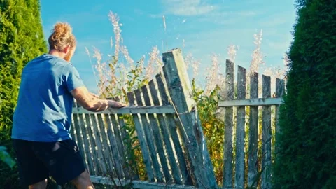 A Man is Removing a Section of an Old Wooden Fence in a Grassy Outdoor Area - Видео 336626023