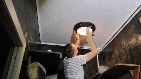Man replaces ceiling light in room with dark walls and wooden shelf during dayti Stock Footage 330188544