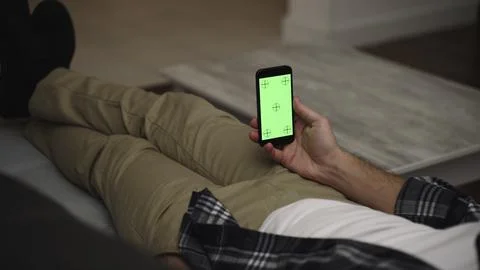 Man resting on a couch at home, using smartphone with green mock-up screen Stock Photos