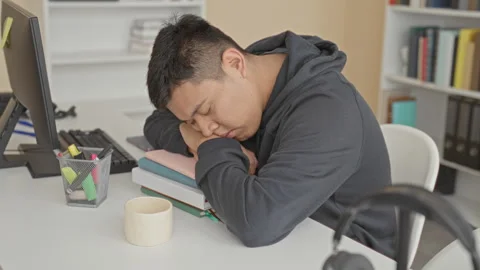 Man rests head on folded hands over stacked books at a desk near computer a.. Stock Footage 347803550