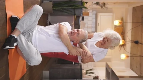 A man of retirement age is sitting on the floor on a fitness mat, and his wife Stock Footage 237912537