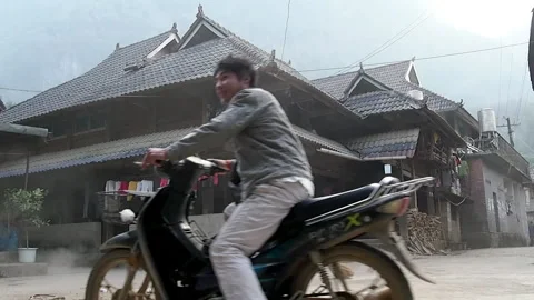A man rides motorcycle through a small Dai village in China’s Yunnan Province.  Stock Footage 158901318