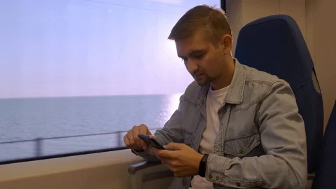 A man rides a train near a window overlooking the sea and uses a phone. Vídeos de archivo 118956070