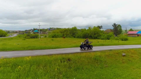 A man riding an ATV on a wet road in a green field. Copter ( drone ) shooting Stock Footage 119521853