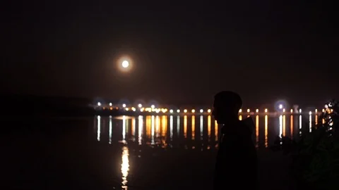 A man by the river in the late evening. Big moon, lights in the background Stock Footage 114631047