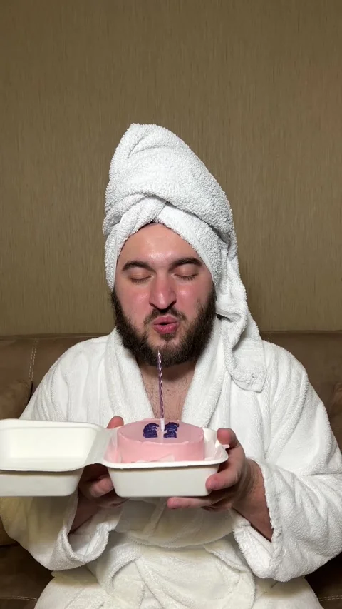 Man in robe, with towel on head blows burning candle Stock Footage 277827314